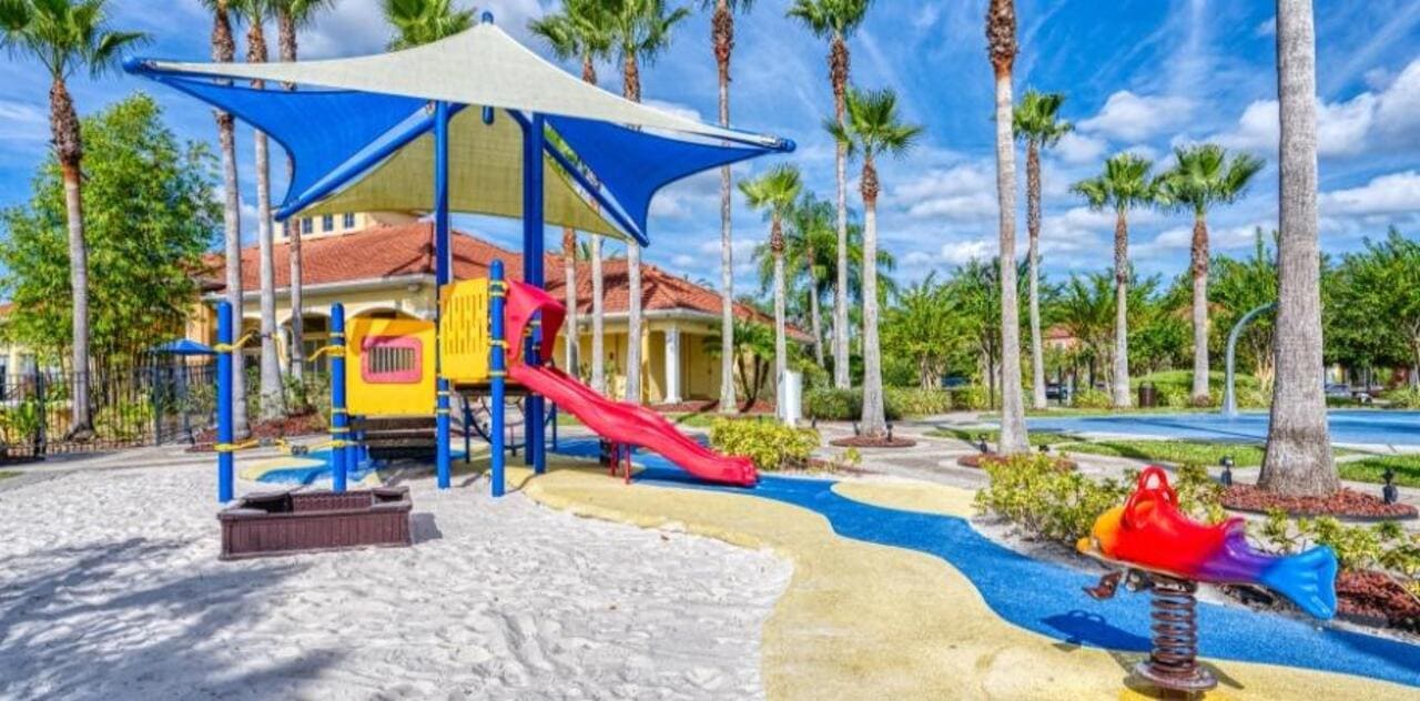 Terra Verde playground with a blue and white shade sail, a red slide, and palms